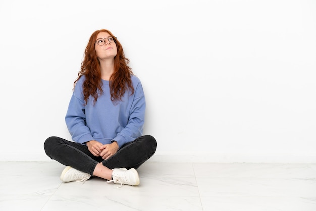 Foto ragazza adolescente rossa seduta sul pavimento isolata su sfondo bianco e alzando lo sguardo