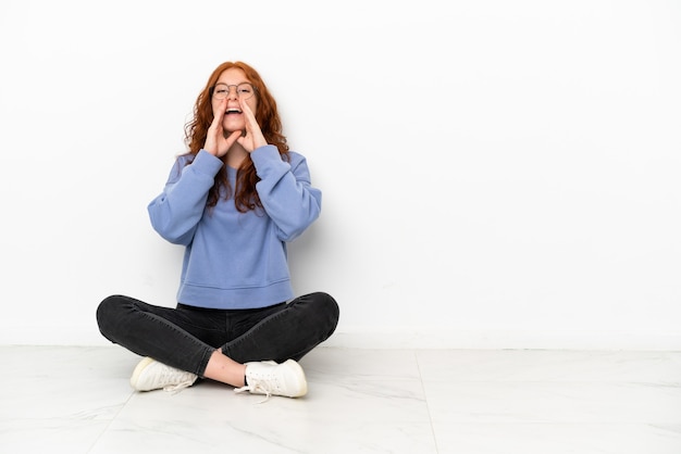 Фото Рыжая девушка-подросток сидит на полу, изолированном на белом фоне, кричит и что-то объявляет