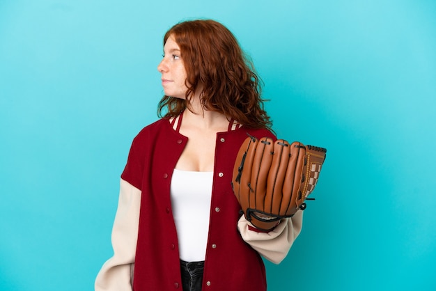 Teenager redhead girl with baseball glove isolated on blue background looking to the side