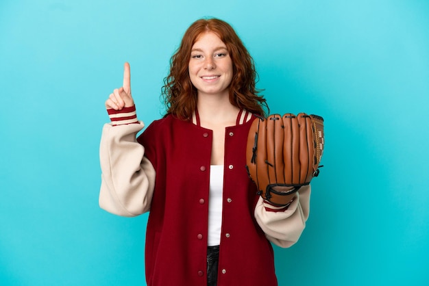 Teenager redhead girl with baseball glove isolated on blue background showing and lifting a finger in sign of the best