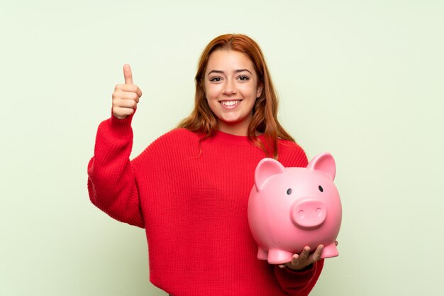 Teenager redhead girl with sweater holding a big piggybank