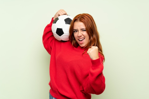 Ragazza della testarossa dell'adolescente con il maglione sopra verde isolato che tiene un pallone da calcio