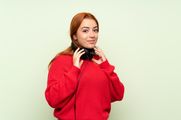 Ragazza rossa adolescente con maglione sopra verde isolato ascoltando musica con le cuffie