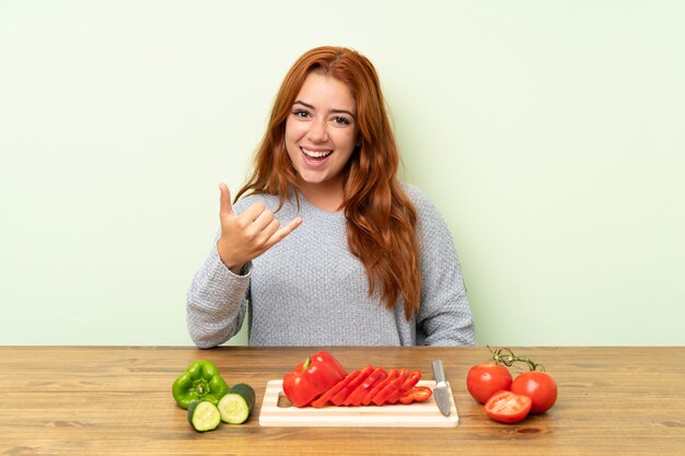 Teenager redhead girl with vegetables in a table making phone gesture