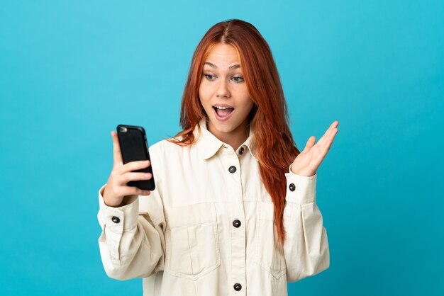 Teenager Russian girl on blue looking at the camera while using the mobile with surprised expression