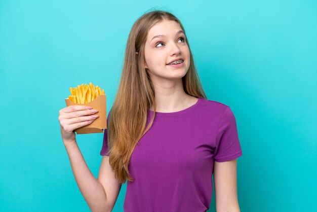 Teenager Russian girl catching french fries isolated on blue background looking up while smiling