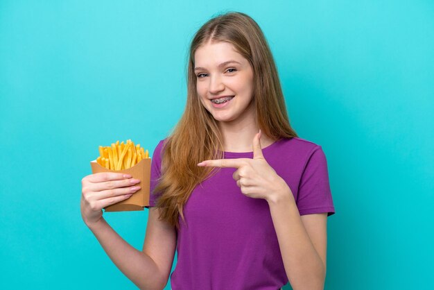 Teenager Russian girl catching french fries isolated on blue background and pointing it