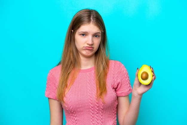 Teenager Russian girl holding an avocado isolated on blue background with sad expression