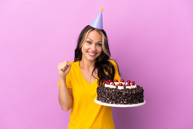 Teenager Russian girl holding birthday cake isolated on purple background celebrating a victory in winner position