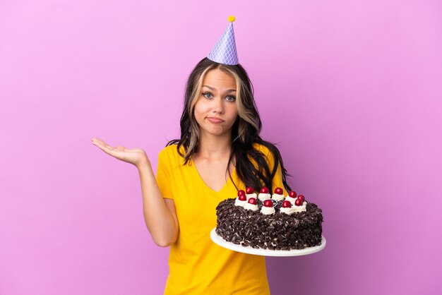 Teenager Russian girl holding birthday cake isolated on purple background having doubts while raising hands