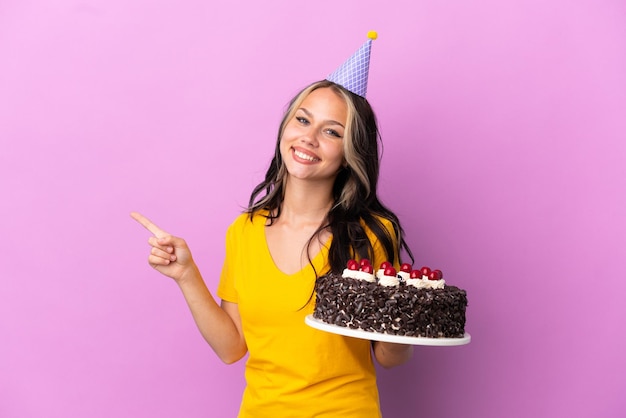 Teenager Russian girl holding birthday cake isolated on purple background pointing finger to the side