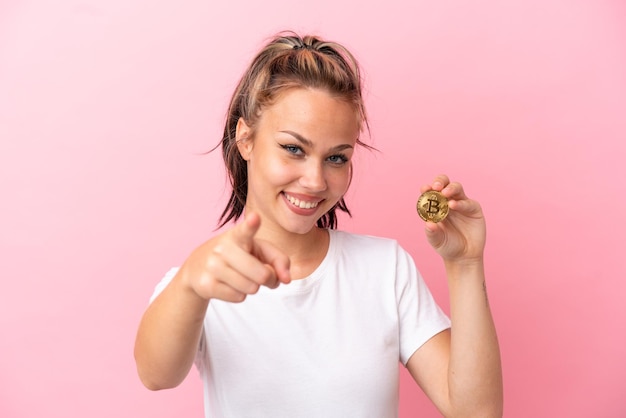 幸せな表情で正面を指すピンクの背景に分離されたビットコインを保持している10代のロシアの女の子