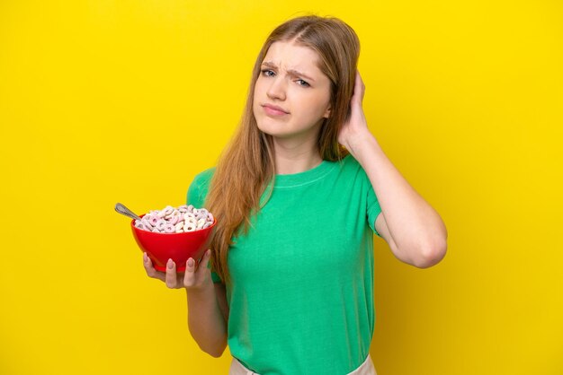 Teenager russian girl holding bowl of cereals isolated on yellow background having doubts