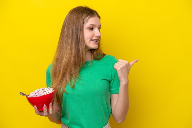 Teenager russian girl holding bowl of cereals isolated on yellow background pointing to the side to present a product