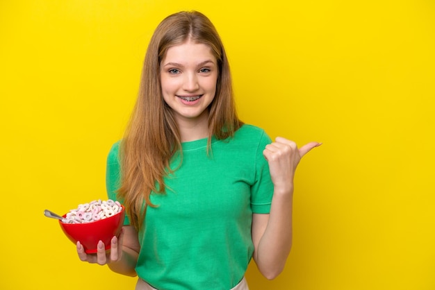 Teenager russian girl holding bowl of cereals isolated on yellow background pointing to the side to present a product