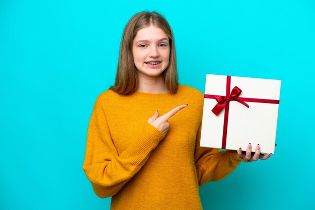 Русская девушка-подросток держит подарок на синем фоне и указывает на него