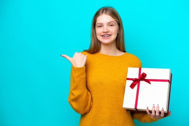 Teenager Russian girl holding a gift isolated on blue background pointing to the side to present a product