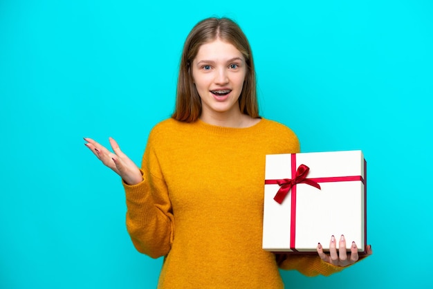 Teenager Russian girl holding a gift isolated on blue background with shocked facial expression