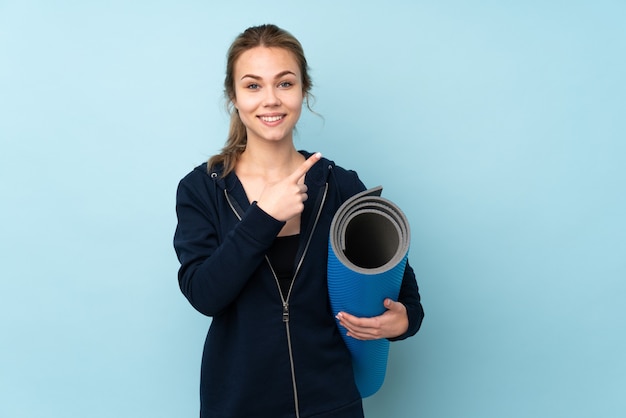 Teenager Russian girl holding mat isolated on blue pointing to the side to present a product
