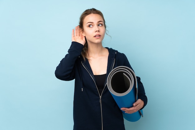 Teenager Russian girl holding mat isolated on blue wall listening to something by putting hand on the ear