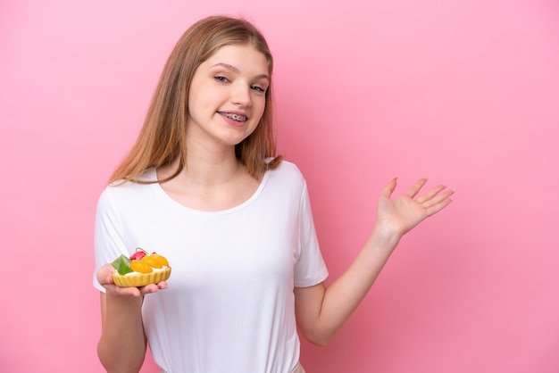 Teenager Russian girl holding a tartlet isolated on pink background extending hands to the side for inviting to come