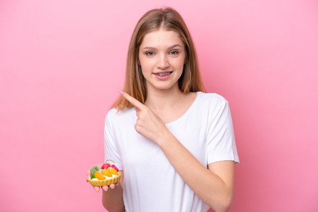 Teenager Russian girl holding a tartlet isolated on pink background pointing to the side to present a product