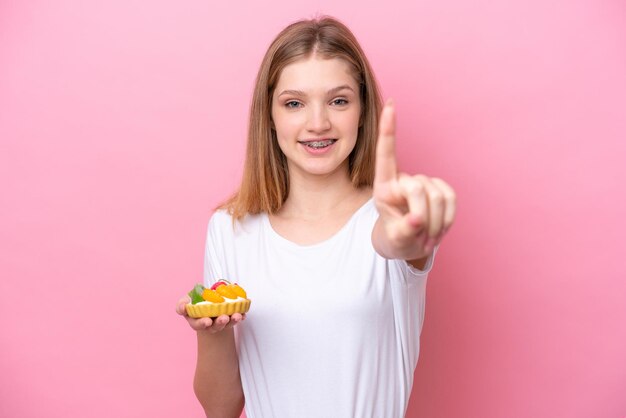 Русская девушка-подросток держит тарталетку на розовом фоне, показывая и поднимая палец