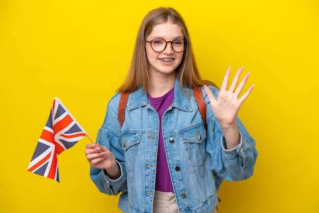 손가락으로 다섯을 세는 노란색 배경에 고립 된 영국 국기를 들고 십 대 러시아 소녀