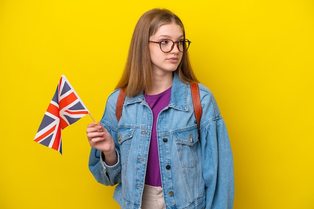 横を見て黄色の背景に分離されたイギリスの旗を保持している10代のロシアの女の子