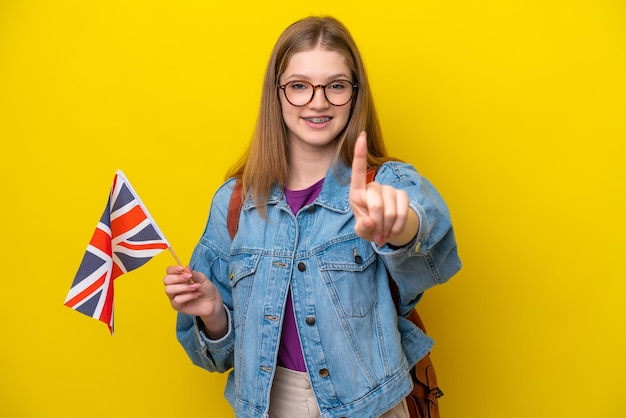 Teenager Russian girl holding an United Kingdom flag isolated on yellow background showing and lifting a finger