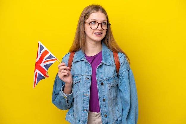 노란색 배경에 격리된 영국 국기를 들고 있는 10대 러시아 소녀가 올려다보는 동안 아이디어를 생각하고 있다
