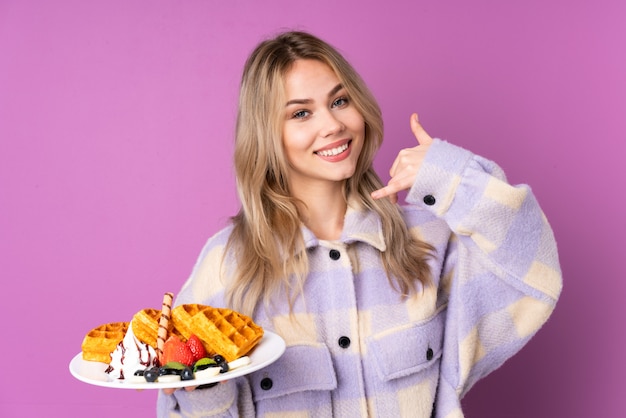 Teenager Russian girl holding waffles on purple wall making phone gesture and pointing front