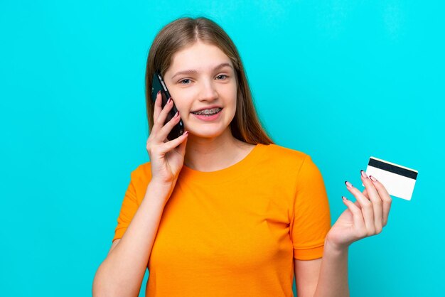 Teenager Russian girl isolated on blue background keeping a conversation with the mobile phone and holding a credit card