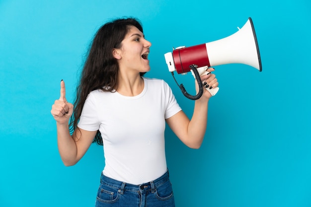 Teenager Russian girl isolated on blue wall shouting through a megaphone to announce something in lateral position