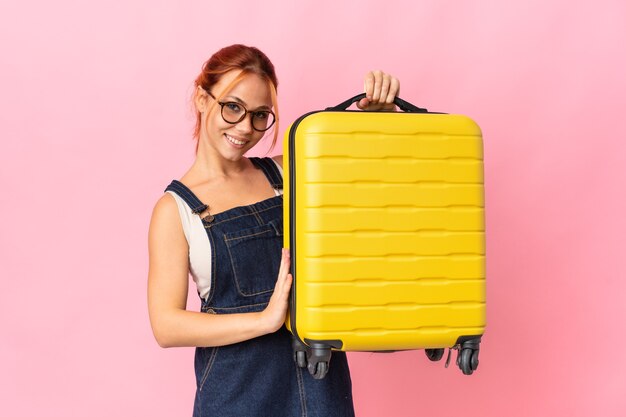 Ragazza russa dell'adolescente isolata sulla parete rosa in vacanza con la valigia di viaggio