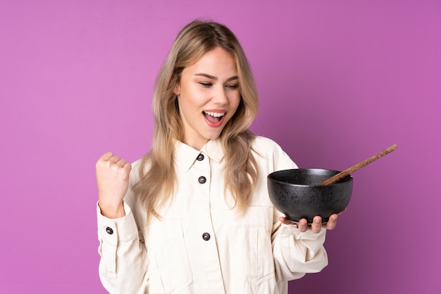 Teenager Russian girl isolated on purple celebrating a victory while holding a bowl of noodles with chopsticks