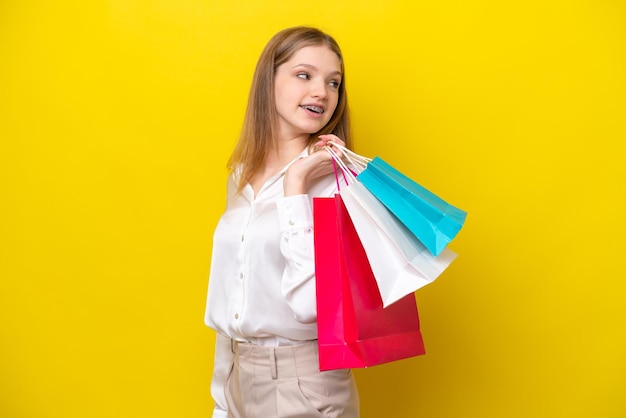 Teenager Russian girl isolated on yellow background holding shopping bags and looking back