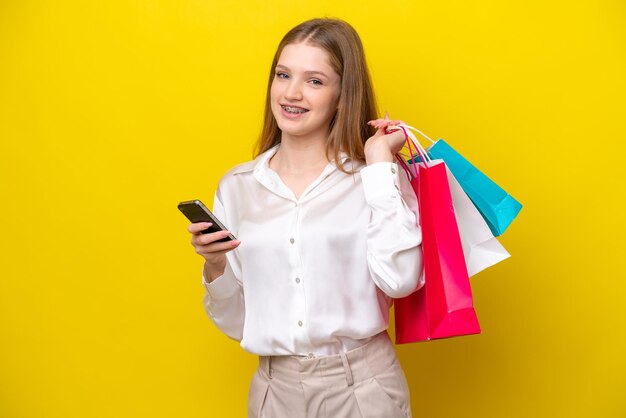 Teenager Russian girl isolated on yellow background holding shopping bags and writing a message with her cell phone to a friend