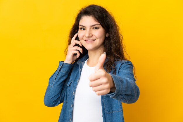 Teenager Russian girl isolated on yellow background keeping a conversation with the mobile while doing thumbs up