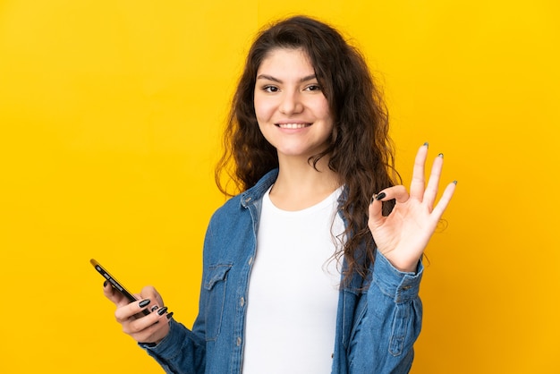 携帯電話を使用してOKサインをしている黄色の背景に分離された10代のロシアの女の子