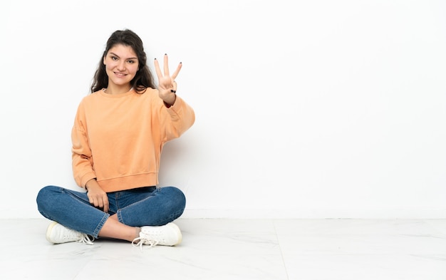 Teenager Russian girl sitting on the floor happy and counting three with fingers