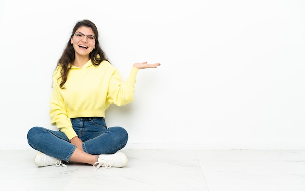 Teenager Russian girl sitting on the floor holding copyspace imaginary on the palm to insert an ad