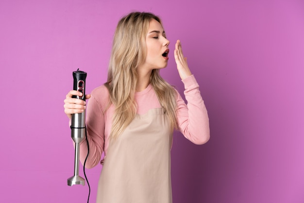 Teenager Russian girl using hand blender isolated on purple wall yawning and covering wide open mouth with hand