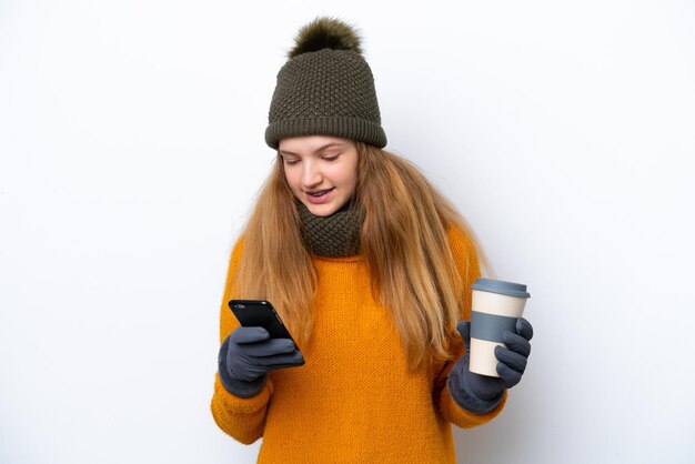 Teenager russian girl wearing winter jacket isolated on white\
background holding coffee to take away and a mobile