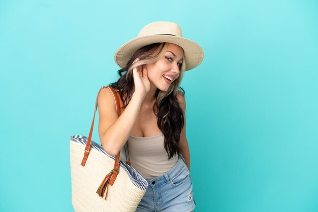 Teenager Russian girl with pamel and beach bag isolated on blue background listening to something by putting hand on the ear