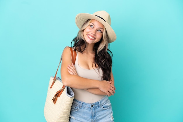 Teenager Russian girl with pamel and beach bag isolated on blue background looking up while smiling