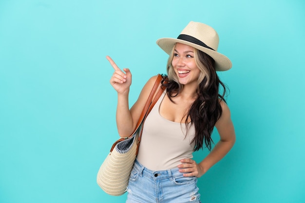 Teenager Russian girl with pamel and beach bag isolated on blue background pointing finger to the side and presenting a product