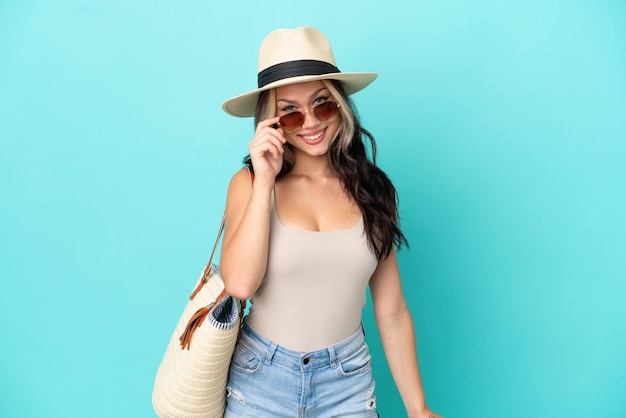 Teenager Russian girl with pamel and beach bag isolated on blue background with glasses and happy