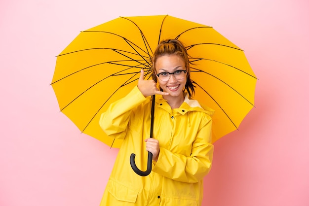 Русская девушка-подросток в непромокаемом пальто и зонтике на розовом фоне делает телефонный жест Перезвони мне