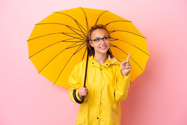 素晴らしいアイデアを指しているピンクの背景に分離された防雨コートと傘を持つ10代のロシアの女の子